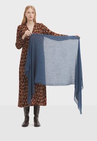 Bufanda ligera azul con una textura delicada, sostenida para mostrar su forma, combinada con un vestido largo marrón estampado y botas negras hasta la rodilla.