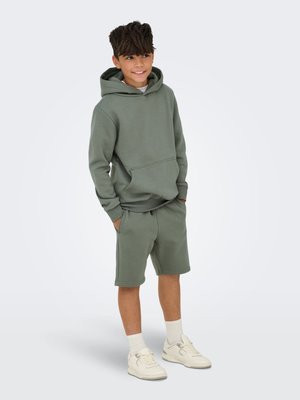 Green hooded sweatshirt and matching shorts, featuring a front pocket and soft texture. Worn with white sneakers and ribbed socks.