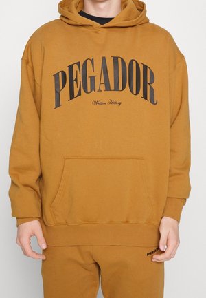 Osoba ubrana w musztardowożółtą bluzę z kapturem i pasujące spodnie, na bluzie napis „PEGADOR Written History” dużymi czarnymi literami na piersi.