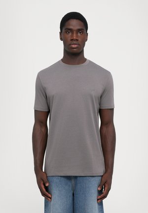 Graues Baumwoll-T-Shirt mit Rundhalsausschnitt und kurzen Ärmeln. Mit einem dezenten Logo auf der Brust. Kombiniert mit hellblauen Jeans.