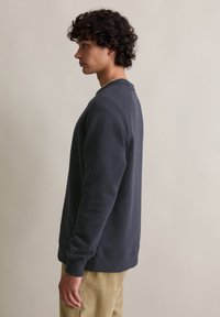 Felpa grigia con colletto rotondo, polsini e vita a costine. Texture liscia. Abbinata a pantaloni in corduroy beige. Vista laterale.