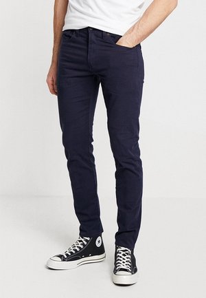 Mężczyzna w ciemnoniebieskich dopasowanych spodniach i czarnych wysokich trampkach z białymi sznurowadłami, stojący z jedną ręką w kieszeni.