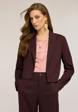 Frau mit langen braunen Haaren trägt einen burgunderroten Anzug mit kurzem Blazer, rosa Oberteil, lange goldene Halskette und große goldene Ohrringe mit Bernstein.