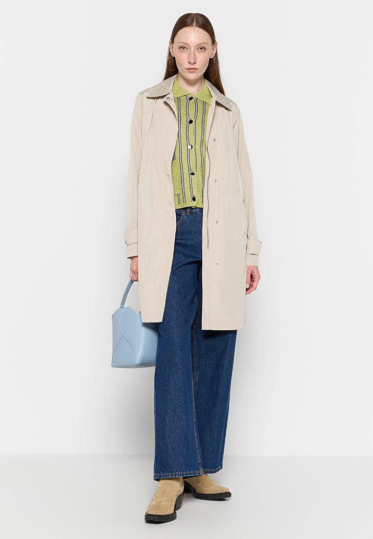 Manteau trench beige avec un col, chemise à rayures vertes à boutons, jeans larges bleus, bottines beige et sac à main bleu clair.