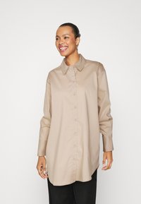 Camicia beige con bottoni, colletto, maniche lunghe con polsini e vestibilità rilassata. Presenta una texture liscia e una leggera lucentezza.