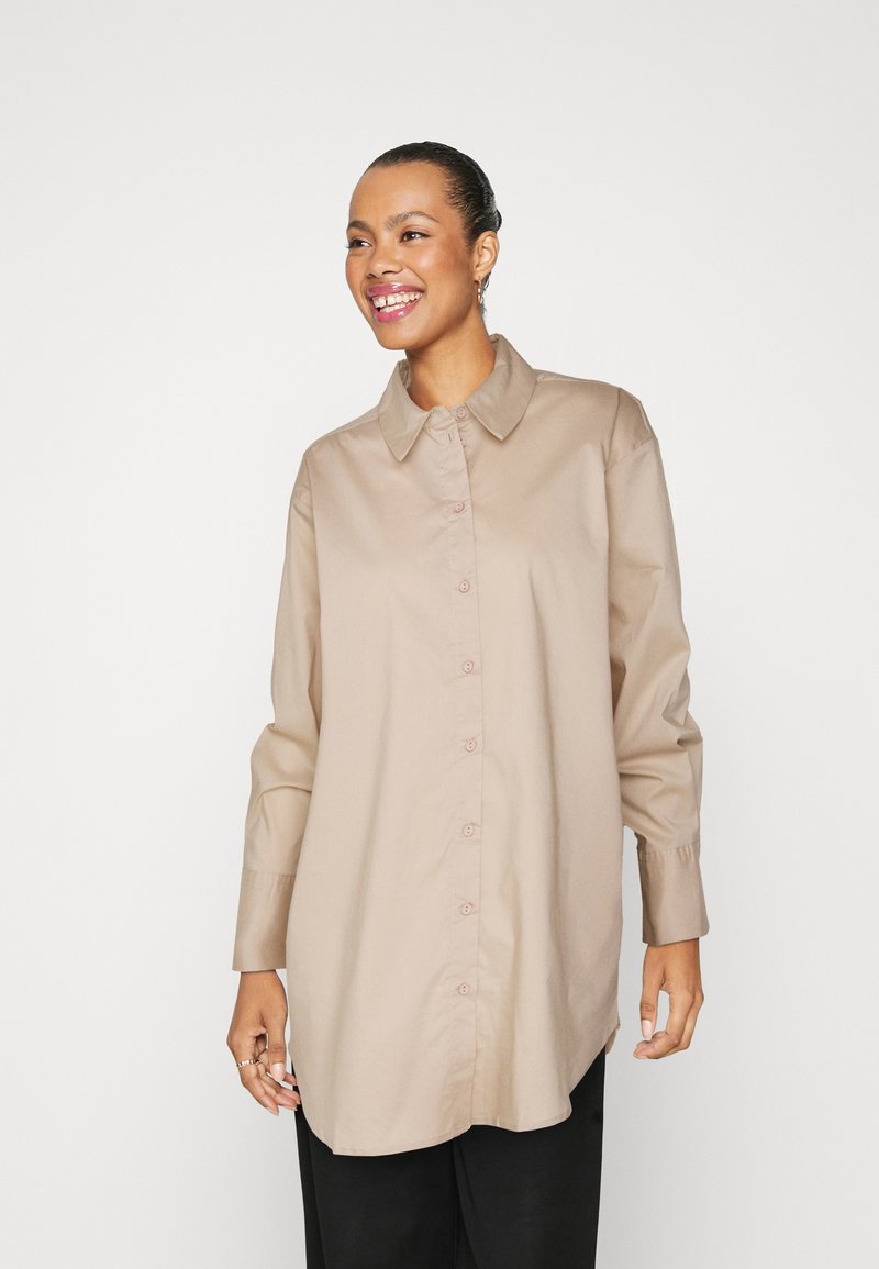 Camicia beige con bottoni, colletto, maniche lunghe con polsini e vestibilità rilassata. Presenta una texture liscia e una leggera lucentezza.