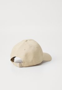 Cappello in cotone beige con visiera curva, fascetta in tessuto regolabile e un bottone in cima. Presenta fori di ventilazione cuciti su entrambi i lati.