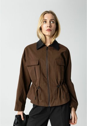 Blonde Frau trägt eine braune Reißverschlussjacke mit schwarzem Kragen und Kordeln und hält eine schwarze strukturierte Clutch in der rechten Hand.