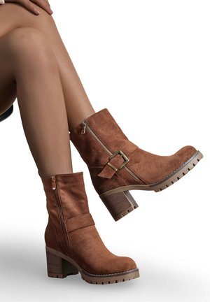 Bottines en suède marron avec une fermeture éclair sur le côté, une lanière à boucle et un large talon. Tige texturée et semelle en caoutchouc avec des détails de crampons.