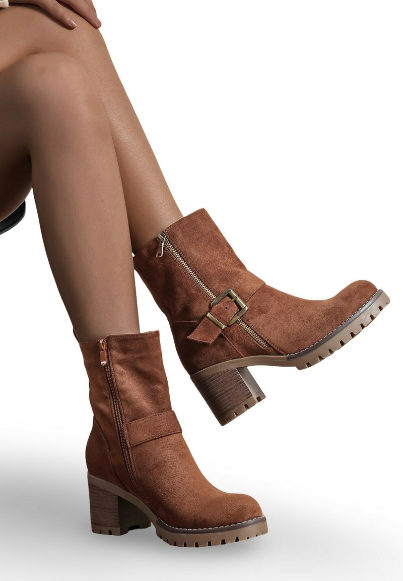 Bottines en suède marron avec une fermeture éclair sur le côté, une lanière à boucle et un large talon. Tige texturée et semelle en caoutchouc avec des détails de crampons.
