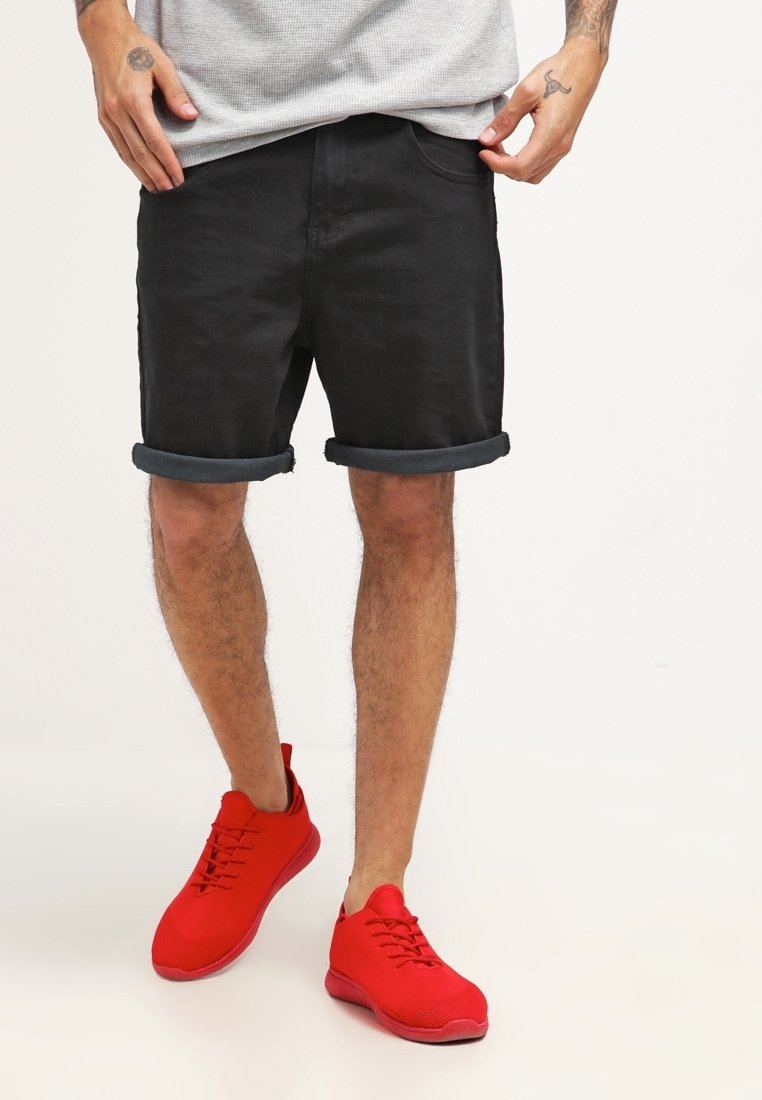 Shorts en denim noir avec revers roulés, associés à des chaussures de sport rouge vif dotées d'une tige texturée et de lacets, visibles sous un haut gris.