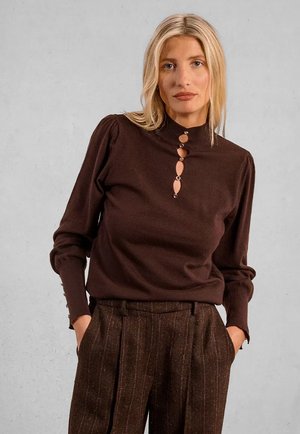 Donna bionda che indossa un top marrone a maniche lunghe con ritagli decorativi e pantaloni a vita alta marroni con texture, mani nelle tasche.
