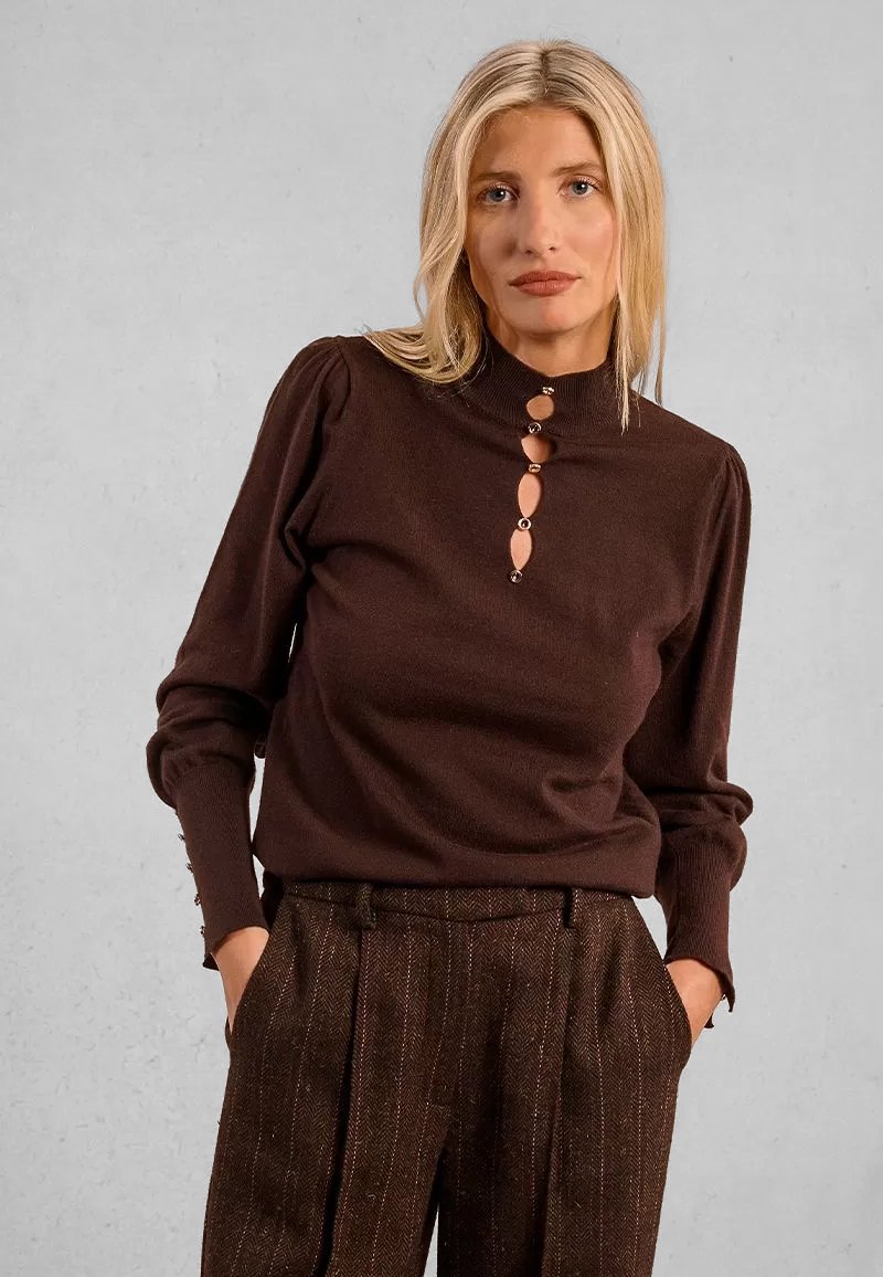 Donna bionda che indossa un top marrone a maniche lunghe con ritagli decorativi e pantaloni a vita alta marroni con texture, mani nelle tasche.