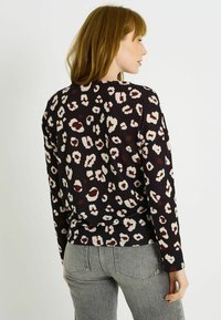 Mujer con cabello rojo que lleva una camiseta de leopardo de mangas largas negras y jeans grises, de lado frente a un fondo plano.