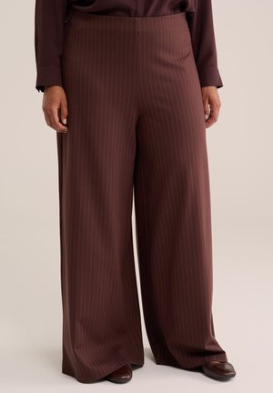 Pantalon marron à jambes larges avec de fines rayures verticales, porté avec des chaussures marron assorties et un haut sombre.