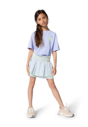 Young girl with long brown hair wearing a light purple t-shirt, blue striped skirt, and beige sneakers standing with legs apart on white background.