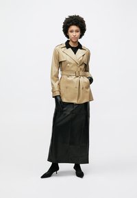 Μπεζ δερμάτινο διπλόμπουτο καμπαρντίνα με ζώνη στη μέση, συνδυασμένο με μαύρη δερμάτινη φούστα μάξι. Μαύρα γάντια και ψηλοτάκουνα ολοκληρώνουν το σύνολο.