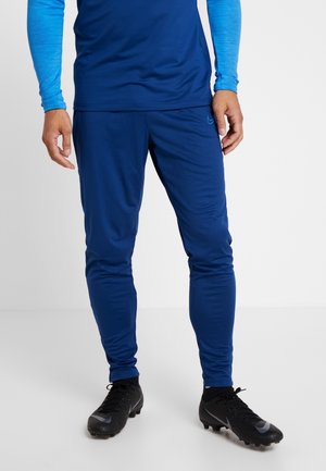 Mężczyzna ubrany w niebieskie sportowe spodnie i pasującą niebieską bluzę z długim rękawem oraz czarne korki, stojący na białej powierzchni na tle jednolitego tła.