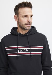 Felpa nera con cappuccio regolabile, caratterizzata da strisce orizzontali bianche e rosse. Logo centrato "FQ1924" in bianco su sfondo striato.