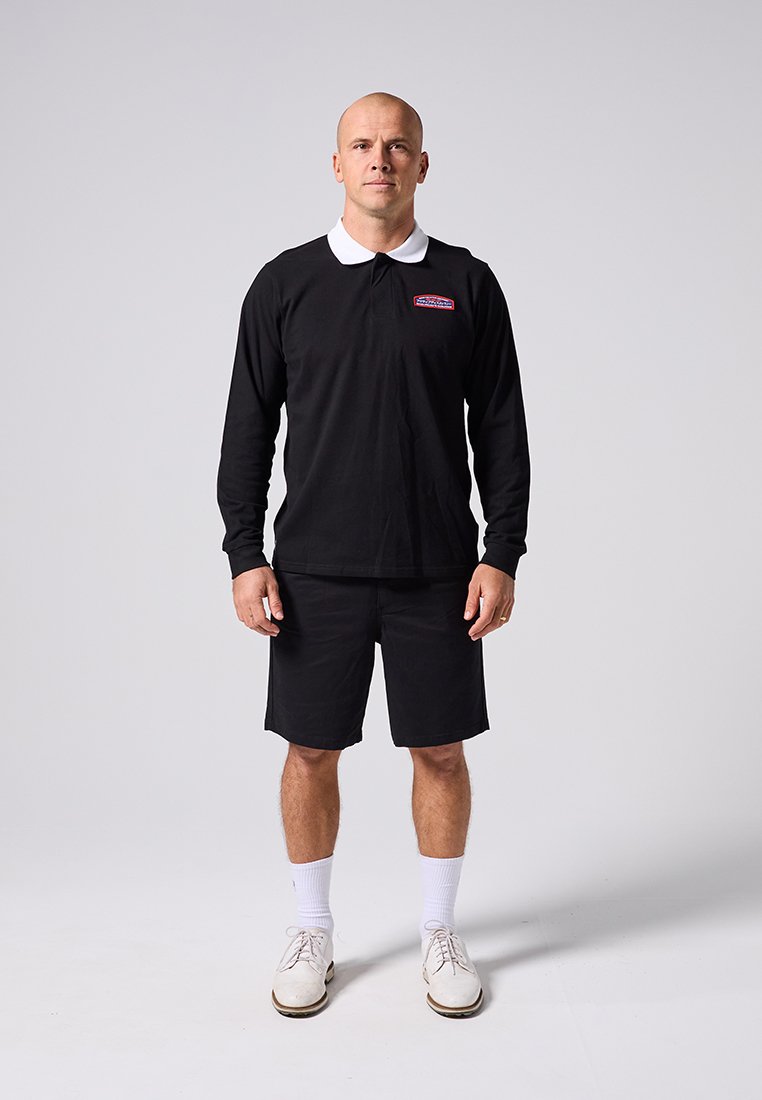 Άνδρας που στέκεται με το πρόσωπο προς τα εμπρός, φορώντας μαύρο πόλο με μακρύ μανίκι, μαύρα σορτς, λευκές κάλτσες και λευκά παπούτσια σε απλό ανοιχτό φόντο.