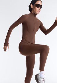 Frau trägt braunes Sportoutfit und große dunkle Sonnenbrille, hebt Knie und Arm in dynamischer Pose vor weißem Hintergrund.