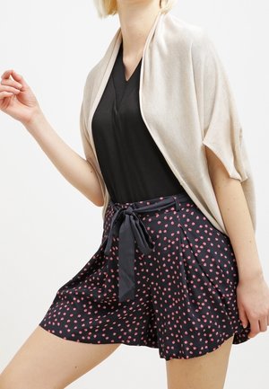 Châle beige clair drapé sur un haut noir à col en V, associé à des shorts navy avec un motif de pois roses et un lien en ruban à la taille.