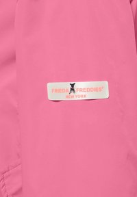 Rosa lättviktsmaterial med slät textur; har en vit etikett med "FRIEDA FREDDIES" och "NEW YORK" samt en liten hundgrafik.