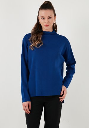 Blauw ribgebreid topje met een hoge hals en lange mouwen, met een ontspannen pasvorm en zijsplitten, gecombineerd met zwarte broek.