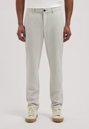 Uomo che indossa pantaloni grigio chiaro a gamba dritta e sneaker bianche, in piedi con le braccia rilassate lungo i fianchi davanti a uno sfondo neutro.