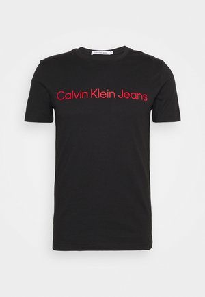 Črna majica s kratkimi rokavi in okroglim izrezom z napisom "Calvin Klein Jeans" v rdeči barvi na prsih.
