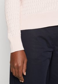 Helle rosa Strickpullover mit strukturiertem Muster, taillierten Bündchen und geripptem Saum. Kombiniert mit dunkelblauen Hosen für einen Kontrast.
