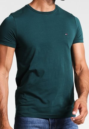 Uomo che indossa una maglietta aderente a maniche corte di colore verde scuro con un piccolo logo sul lato sinistro del petto e jeans blu.