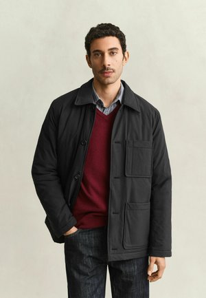 Mann trägt eine schwarze Jacke über einem roten Pullover und einem grauen Hemd, steht mit einer Hand in der Tasche vor einem schlichten hellen Hintergrund.
