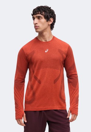 Junger Mann trägt ein eng anliegendes rotes langärmeliges Sportshirt mit Mustern und blickt nach links vor einem schlichten hellen Hintergrund.