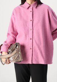 Pink wool shirt with a spread collar, button front, and loose fit. The model holds a metallic silver handbag with chain accents.