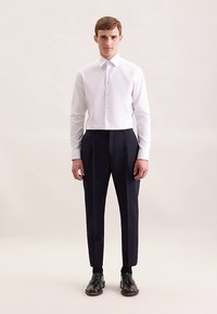 Camicia bianca con bottoni dal design aderente, abbinata a pantaloni scuri a pieghe. L'outfit è completato da scarpe in pelle nere e calze.