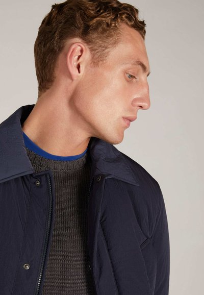 Veste légère navy à finition texturée, dotée d'un col à boutons et d'une fermeture éclair à l'avant, portée par-dessus un pull en maille côtelée marron.