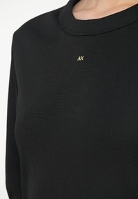 Κοντινή λήψη ενός ατόμου που φοράει ένα μαύρο ριμπ μπλουζάκι με μακριά μανίκια και ένα μικρό χρυσό λογότυπο "AX" κεντραρισμένο κάτω από τον στρογγυλό λαιμό.