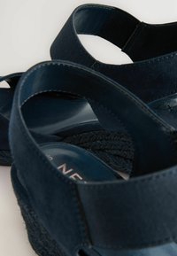 Marineblå kilehæle med bløde ruskindsremme, elastiske detaljer og en struktureret jutesål; glat inderside med logo-detalje.