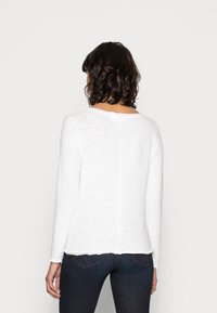 Camisa blanca de manga larga con cuello redondeado, que presenta una textura suave y una costura trasera recta; combinada con jeans oscuros.