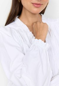 Hvid bluse med rynkede detaljer, elastiske manchetter og en blød tekstur. Nærbillede af halsudskæringen og hånden hviler mod stoffet.