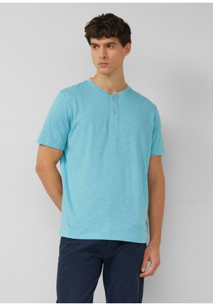 Mann trägt ein kurzärmeliges hellblaues Henley-Shirt und dunkelblaue Hose, steht mit einer Hand hinter dem Rücken vor einem schlichten grauen Hintergrund.