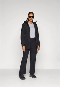 Czarna wodoodporna kurtka z zamkiem błyskawicznym z przodu, kieszeniami i kapturem, zestawiona z szarą koszulką, czarnymi spodniami bojówkami i butami trekkingowymi.