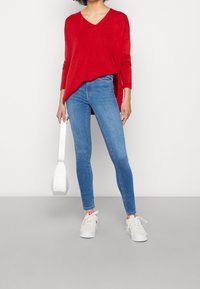 Suéter rojo de manga larga con escote en V combinado con jeans skinny azules. Un bolso blanco tipo cubo y zapatillas blancas con detalles en rosa completan el atuendo.