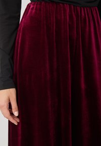 Fustă de catifea bordeaux cu un design fluid și talie elastică, având o textură moale și o cădere netedă. O mână atinge ușor materialul.