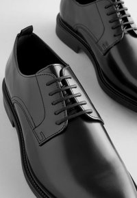 Chaussures habillées en cuir noir avec une finition brillante, présentant un bout rond, six œillets et des lacets noirs. Points de couture remarquables le long des bords.