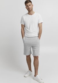 Bărbat stând cu mâinile în buzunare, purtând un tricou alb, pantaloni scurți gri deschis și adidași albi, pe un fundal simplu de culoare deschisă.