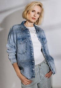 Světle modrá denimová košile s knoflíkovým zapínáním, dvěma kapsami na prsní části a ohrnutými rukávy, spárovaná s bílým tričkem a pruhovanými kalhotami.