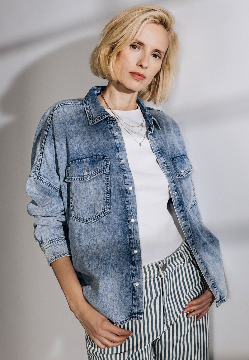Camicia in denim azzurro chiaro con bottone frontale, due tasche sul petto e maniche arrotolate, abbinata a una t-shirt bianca e pantaloni a righe.