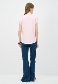 Camicetta rosa chiaro a maniche corte con colletto, abbinata a jeans in denim blu scuro a zampa d'elefante con bottoni laterali e leggere aperture all'orlo.